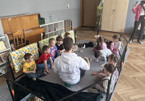 Zdjęcie przedstawia dzieci i mężczyznę siedzących w zagrodzie z królikami.