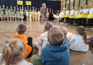 sala gimnastyczna, przed dziećmi stoi kobieta, w ręku trzyma mikrofon