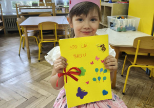 Na zdjęciu widzimy dziewczynkę, która ma na głowie koronę oraz w ręku trzyma upominek w formie książeczki z rysunkami.