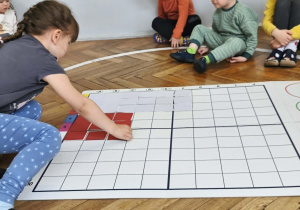 Na zdjęciu widzimy dziewczynkę, która kładzie czerwony kartonik na odpowiednim polu na macie do kodowania.