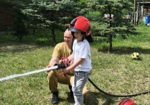 Na zdjęciu widzimy dziecko, które trzyma wąż strażacki i polewa wodę na słupki.