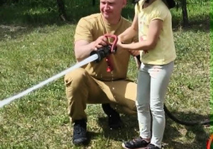 Na zdjęciu widzimy dziecko, które trzyma wąż strażacki i polewa wodę na słupki.