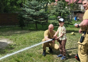 Na zdjęciu widzimy dziecko, które trzyma wąż strażacki i polewa wodę na słupki.