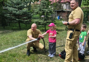 Na zdjęciu widzimy dziecko, które trzyma wąż strażacki i polewa wodę na słupki.