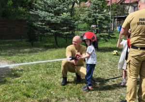 Na zdjęciu widzimy dziecko, które trzyma wąż strażacki i polewa wodę na słupki.