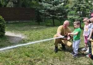 Na zdjęciu widzimy dziecko, które trzyma wąż strażacki i polewa wodę na słupki.