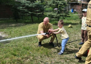 Na zdjęciu widzimy dziecko, które trzyma wąż strażacki i polewa wodę na słupki.