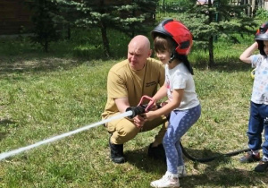 Na zdjęciu widzimy dziecko, które trzyma wąż strażacki i polewa wodę na słupki.