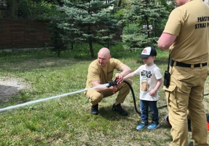 Na zdjęciu widzimy dziecko, które trzyma wąż strażacki i polewa wodę na słupki.