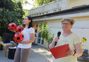 Zdjęcie przedstawia dwie kobiety: jedna przemawia przez mikrofon a druga trzyma trzy piłki.