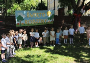 Zdjęcie przedstawia dzieci stojące w półkolu, na tle banera, podczas występu.