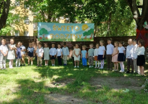 Zdjęcie przedstawia dzieci stojące w półkolu, na tle banera, podczas występu.