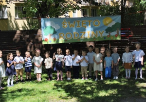Zdjęcie przedstawia dzieci stojące w półkolu, na tle banera, podczas występu.