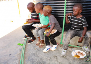 Zdjęcie przedstawia kenińskie dzieci siedzące na niskim murku spożywające posiłek.