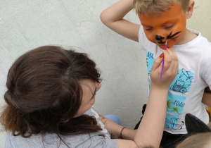 Zdjęcie przedstawia nauczycielkę malującą chłopcu twarz na pomarańczowo.