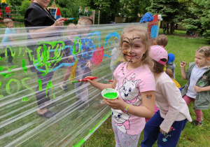 Zdjęcie przedstawia dziewczynkę malującą farbą po folii. Dziewczynka uśmiecha się do obiektywu.