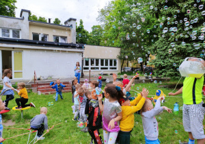 Zdjęcie przedstawia dzieci tańczące w ogrodzie przedszkolnym. W górze fruwają bańki.