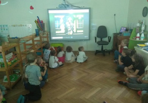 Zdjęcie przedstawia grupę dzieci siedzącą przed tablicą multimedialną, oglądającą akcję strażaków w czasie powodzi.