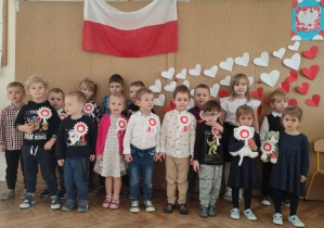 w tle wisi flaga Polski, pod nią z cers w kolorach białych i czerwonych ułożona jest flaga, w prawym górnym rogu widać godło Polski wykonane przez dziecko. Przed flagami stoją dzieci, które do bluzek mają mają dopięte kotyliony w barwach biało czerwonych