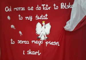 na czerwonym tle znajduje się orzeł biały oraz napis Od morza aż do Tatr to Polska, to mój świat, to serca mego pieśń i skarb