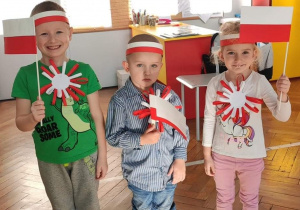 troje dzieci stoją dzieci trzymając w ręku flagi Polski, do bluzek mają dopięte kotyliony w barwach biało czerwonych a na głowach biało czerwone opaskiopaski