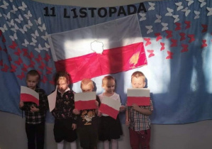 na niebieskim tle jest napis 11 listopada na środku jest flaga Polski po bokach flagi znajdują się białe i czerwone motyle. Przed napisem stoją dzieci z flagami Polski w ręku
