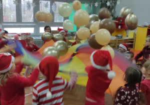 Zdjęcie przedstawia dzieci ubrane świątecznie, które stoją w kole i podrzucają balony na chuście animacyjnej.