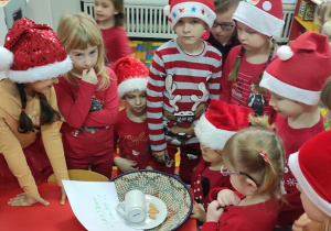 Zdjęcie przedstawia dzieci ubrane w świąteczne akcenty, które są zebrane wokół talerzyka z pustym kubeczkiem, pokruszonymi ciasteczkami i listem od Mikołaja.