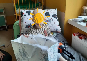 Na zdjęciu widać małe dziecko siedzące na szpitalnym łóżku, które trzyma torbę z prezentami