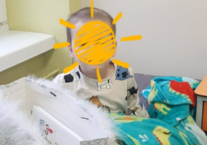 Zdjęcie przedstawia dziecko siedzące na łóżku szpitalnym z torbą z prezentami