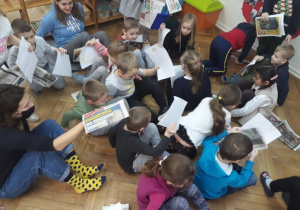 Zdjęcie przedstawia dużą grupę dzieci, która siedząc na gazecie przemieszcza się po sali