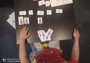 Chłopiec siedzi przy stole, na którym leżą pomoce dydaktyczne: cyfry, znaki i liczmany w postaci obrazków przedstawiająycch SUPER MARIO