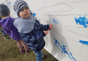 Zdjęcie przedstawia chłopca malującego niebieską farbą po kartonie rozłożonym w ogrodzie przedszkolnym.
