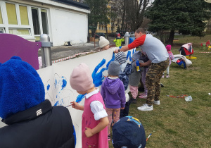 Zdjęcie przedstawia dzieci i nauczyciela malujących farbami po kartonie rozłożonym w ogrodzie przedszkolnym.