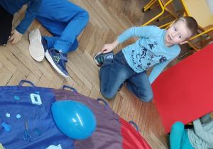 Zdjęcie przedstawia dzieci siedzące wokół chusty animacyjnej. Na niebieskim kolorze chusty położone są różne niebieskie przedmioty.