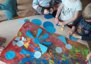Zdjęcie przedstawia dzieci wykonujące plakat. Dzieci wykonują niebieskie kwiaty, które przyklejają na kolorowy karton.
