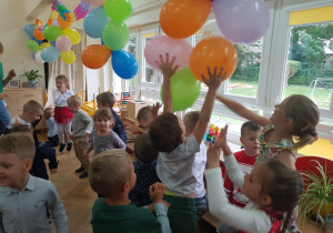 Zdjęcie przedstawia tańczące dzieci. Niektóre z nich próbują dosięgnąć wiszących balonów.