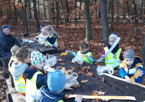 Zdjęcie przedstawia grupę dzieci, które tworzą przy stolikach korony dzieci lasu z materiału które w nim znalazły i zebrały