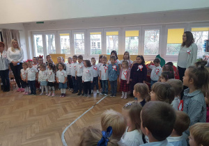 Zdjęcie przedstawia salę wypełnioną dziećmi oraz nauczycielkami . Wszyscy są ubrani w odświętne stroje.