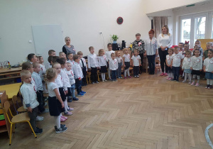Zdjęcie przedstawia salę przedszkolną w której odświętnie ubrane dzieci oraz pracowanicy placówki odśpiewują hymn „Mazurka Dąbrowskiego"..