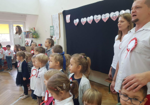 Zdjęcie przedstawia dziecśpiewających i oraz nauczycieli przedszkola hymn Polski. Na drewnianych drzwiach sali widnieje dekoracja z napisem „11 LISTOPADA".