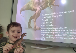 Zdjęcie przedstawia chłopca stojącego z dinozaurem przed tablicą multimedialną