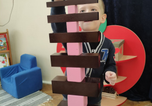 Zdjęcie przedstawia chłopca układającego wysoką konstrukcję z wykorzystaniem brązowych schodów oraz różowej wieży.