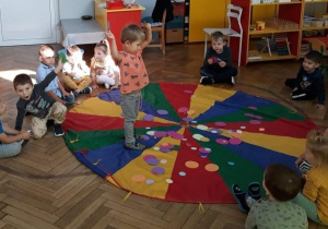 Zdjęcie przedstawia dzieci siedzące wokół chusty animacyjnej na której leżą kółka z papieru. Jeden z chłopców spaceruje po chuście.